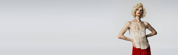 Королева трансвеститов в блондинистом парике позирует в красной юбке и кремовом корсете на сером фоне.