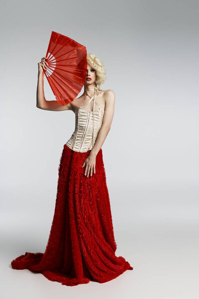 Королева трансвеститов в светлом парике позирует с красным вентилятором, в красной юбке и с белым корсетом..