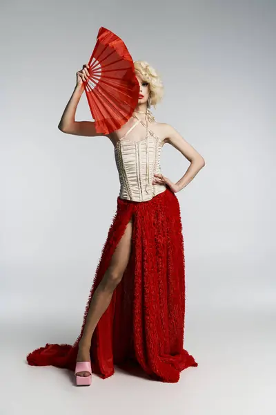 Stock image A drag queen in a blonde wig, red skirt, and white corset poses with a red fan in front of a gray background.