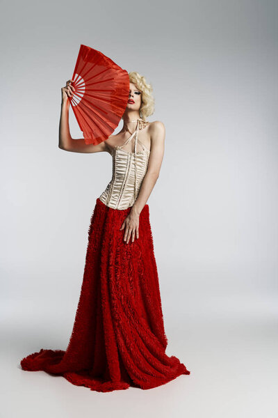 Королева-трансвестит в красной юбке и корсете позирует с ручным вентилятором перед серым фоном.