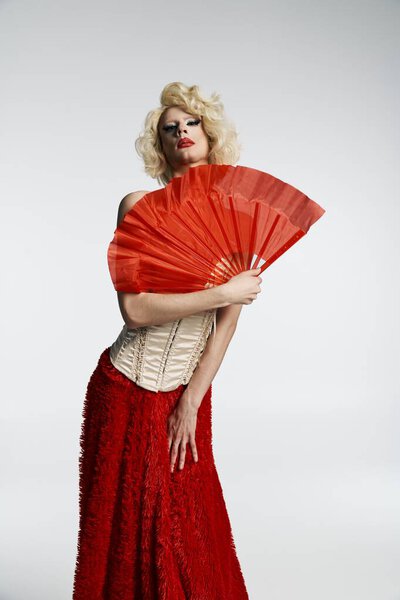 Королева трансвеститов в светлом парике, красной юбке и корсете держит красный вентилятор перед серым фоном.