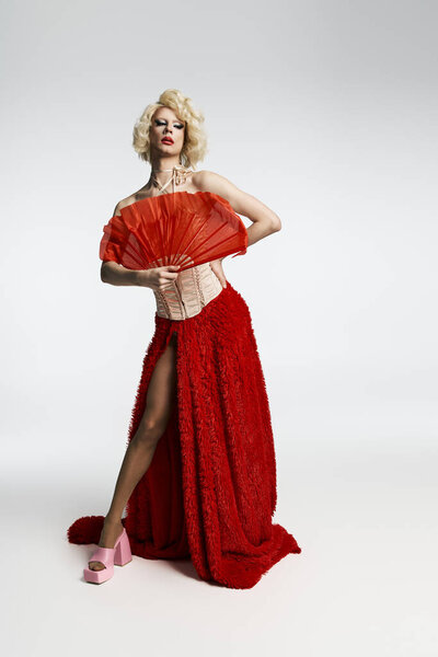 Королева-трансвестит с светлыми волосами и смелым макияжем позирует в красной юбке и корсете, держа в руках красный веер.