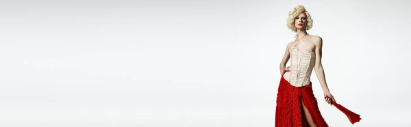 Королева трансвеститов позирует в светлом парике и красной юбке с белым корсетом на сером фоне.