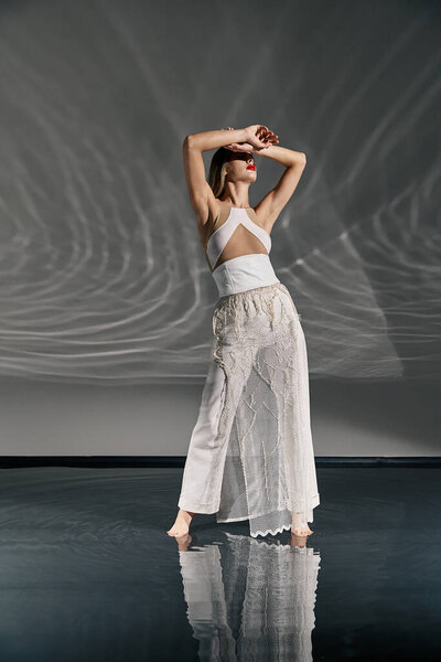 A young woman in a white outfit poses on a reflective floor, with a gray background behind her.