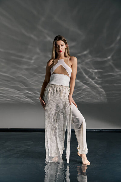 A young woman in a white outfit poses on a grey background, with a reflective surface that makes her appear to be walking on water.