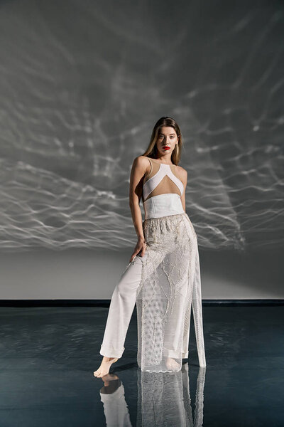A young woman in a white outfit poses on a grey background with a white, wave-like pattern projected onto the wall.