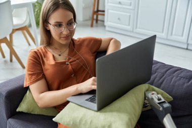 A woman sits on a couch, using a laptop with a prosthetic leg resting on the cushion beside her. clipart