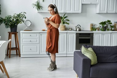 A woman with a prosthetic leg stands in a modern kitchen, using her phone. clipart