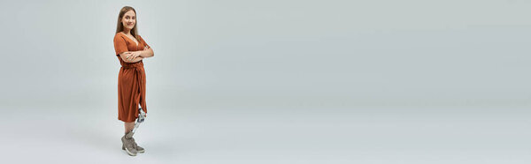 A woman with a prosthetic leg stands confidently with arms crossed, wearing a copper dress.