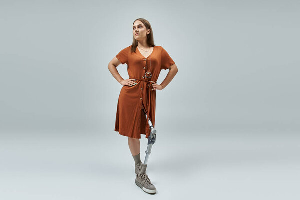 Уверенная женщина стоит с руками на бедрах, в коричневом платье и протезной ноге..
