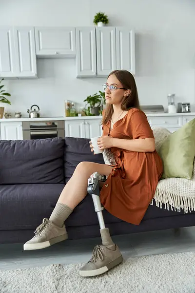 stock image A woman sits on a couch in a living room, looking out the window, holding a cup in her hand.