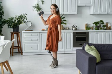 Protez bacaklı bir kadın modern bir mutfakta duruyor, müzik dinliyor ve gülümsüyor..