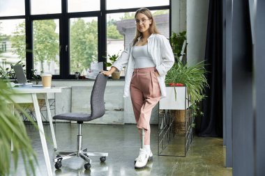 Woman confidently stands with prosthetic leg in modern office setting, leaning on chair clipart