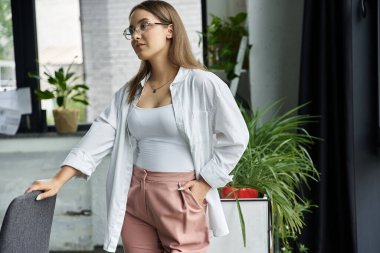 Beyaz gömlekli ve pembe pantolonlu bir kadın modern ofis ortamında sandalyeye yaslanıyor..