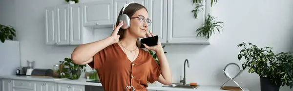 stock image A woman stands in a kitchen, wearing headphones and talking on her phone.