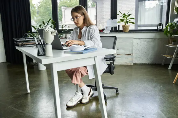 stock image A woman with a prosthetic leg works on her laptop at her desk.