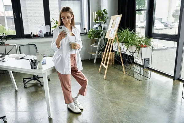 stock image Woman in white shirt and pink pants, leaning against desk, checks phone and holds coffee cup