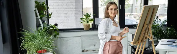 stock image A woman in a white shirt and pink pants stands confidently in front of an easel, ready to create.