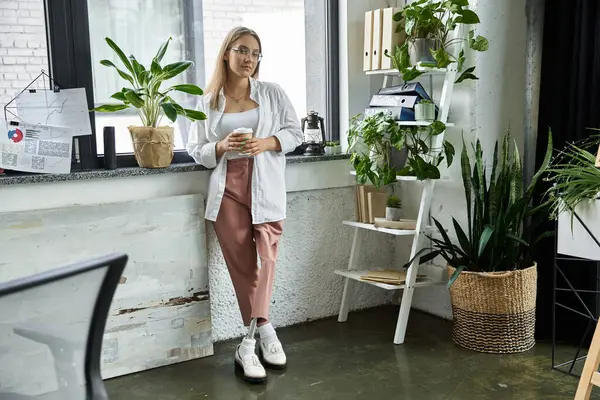 stock image A woman stands by a window, enjoying her coffee and looking out at the city.