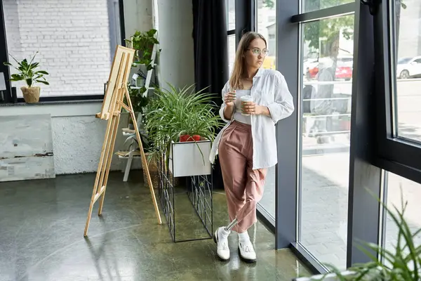 stock image A woman with a prosthetic leg leans against a large window in a studio, holding a coffee cup.