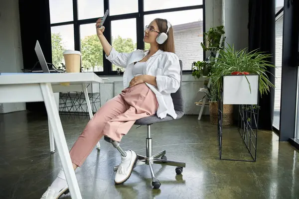 stock image A young woman in an office setting smiles as she takes a selfie while wearing headphones.