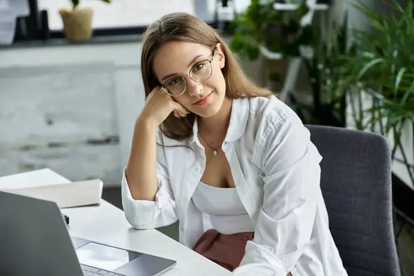 stock image A woman in a white shirt sits at a desk, resting her chin on her hand.