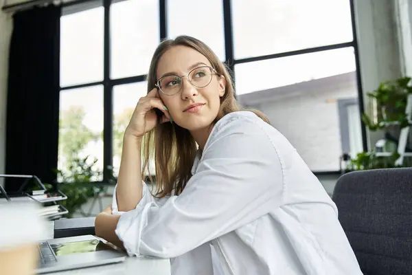 Stock image A woman wearing glasses sits at her desk, thoughtfully looking away from the camera.