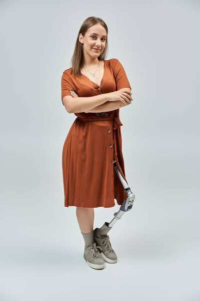 Женщина с протезной ногой уверенно стоит в коричневом платье со скрещенными руками.