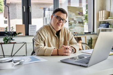 A young man with Down syndrome sits at a desk, laptop open, and looks thoughtfully at the camera. clipart