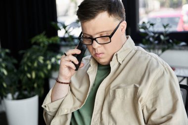 Down sendromlu, gözlüklü, bej gömlekli genç bir adam ev içi telefon görüşmesi yapıyor.