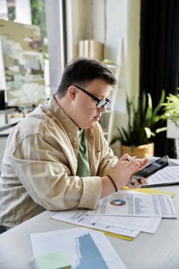 A young man with Down syndrome works on his smartphone at his desk. clipart