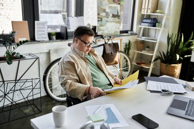 A young man with Down syndrome sits at a desk in an office, reviewing documents. clipart