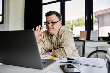 A young man with Down syndrome sits at a desk, waving to someone on a video call. clipart