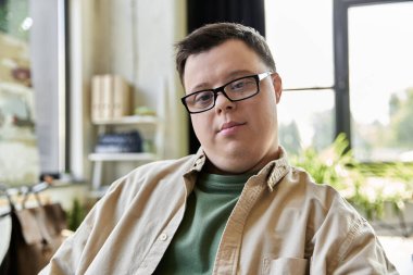 A man with Down syndrome, wearing beige shirt and glasses, looks at camera in a room clipart