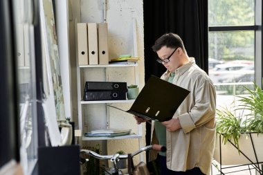 Down sendromlu genç bir adam kitaplığın yanında duruyor ve belgeleri inceliyor..