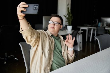 Down sendromlu genç bir adam ofiste selfie çekerken gülümseyip el sallıyor..