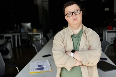 Down sendromlu bir adam kollarını kavuşturmuş bir ofis ortamında kendinden emin bir şekilde duruyor..