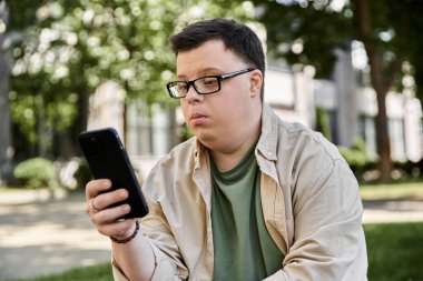 Down sendromlu bir adam parkta oturuyor ve akıllı telefonuna odaklanıyor..