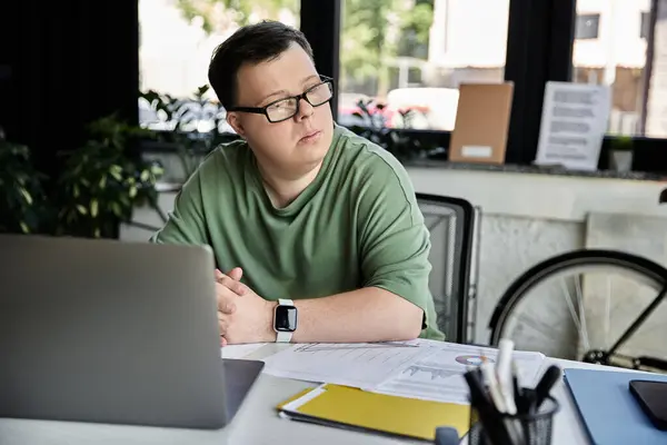 Stock image A young man with Down syndrome in glasses sits at a desk, thoughtfully looking away from his laptop.