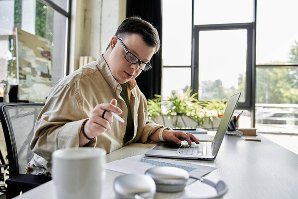 A young man with Down syndrome sits at a desk, working on his laptop and holding a pen.