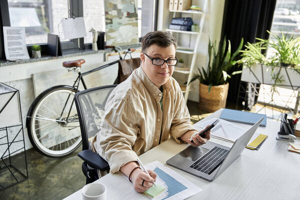 Молодой человек с синдромом Дауна сидит за столом, работает над ноутбуком и графиком, с велосипедом на заднем плане.