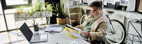 A young man with Down syndrome sits at a desk reviewing documents, while a bicycle rests against the wall behind him.