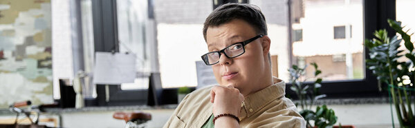 A young man with Down syndrome sits thoughtfully in a room, looking out the window.