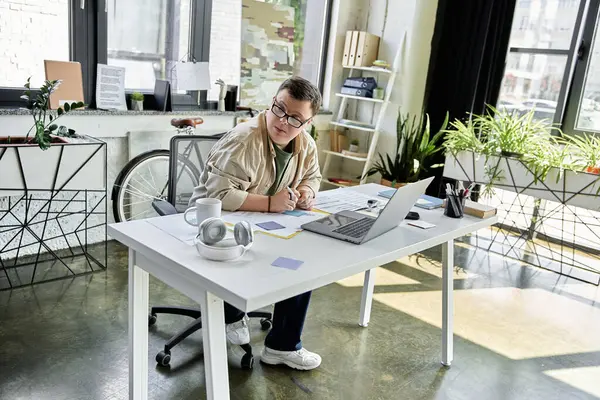 stock image A young man with Down syndrome sits at a desk in a modern office, working on a laptop and writing on a notepad.