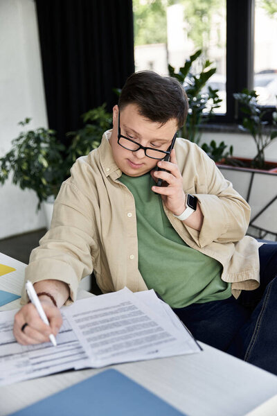A young man with Down syndrome sits at a desk, taking notes while on a phone call.