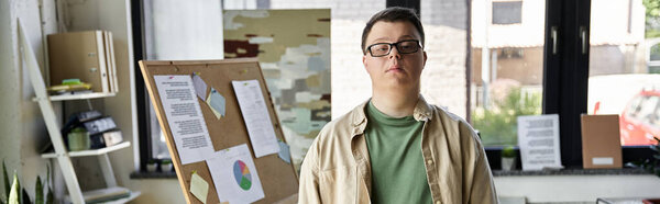 Молодой человек с синдромом Дауна уверенно стоит перед доской объявлений в офисе.