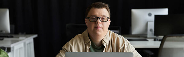 A young man with Down syndrome sits at a desk in an office, working on his laptop.