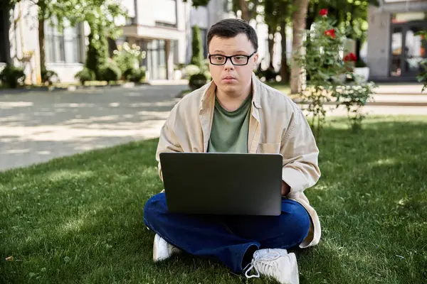 stock image A young man with Down syndrome sits on the grass, focused on his laptop.