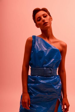 Bir kadın stüdyoda duruyor, geri dönüşümlü plastikten yapılmış mavi bir elbise giyiyor..