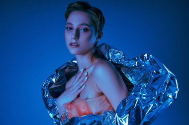 Kadın, parlak gümüş kumaşlı, mavi ışıkla aydınlatılmış sürdürülebilir moda giysileri giyer.
